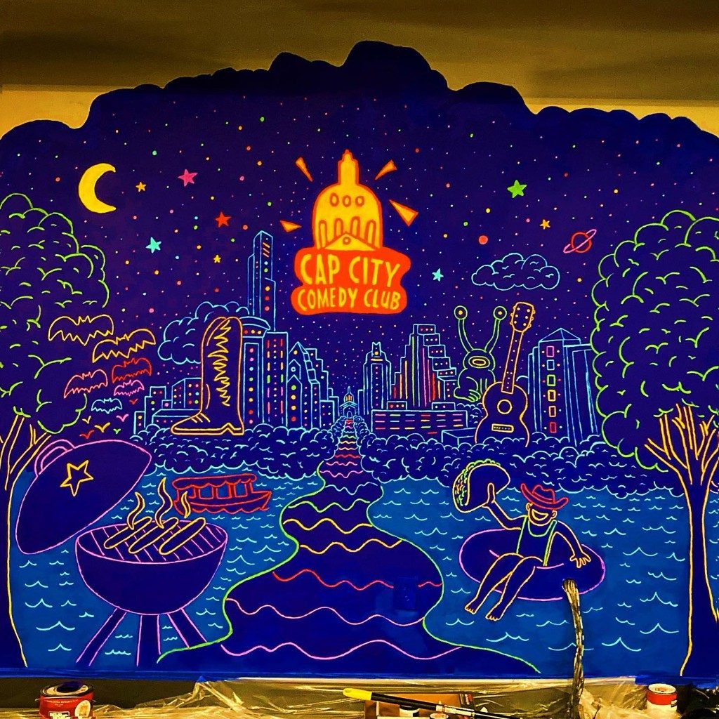 A fluorescent mural of an Austin night: the capitol, a skyline, a river, a grill, and a cowboy in an inner tube. The sign reads Cap City Comedy Club.